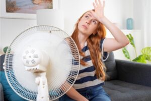 woman feeling heat exhaustion in front of a house fan while looking up at a broken AC