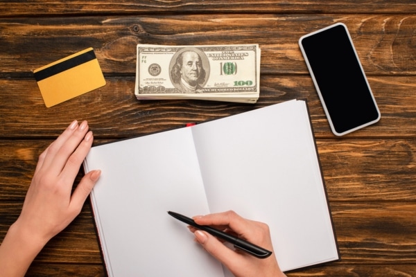 cropped view of a woman's hand about to write on a notebook with credit card, cash, and mobile phone around depicting cost considerations